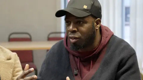 Anthony King, wearing a dark red hooded top and black sweater and a black baseball cap, sits in front of a table and chairs. 