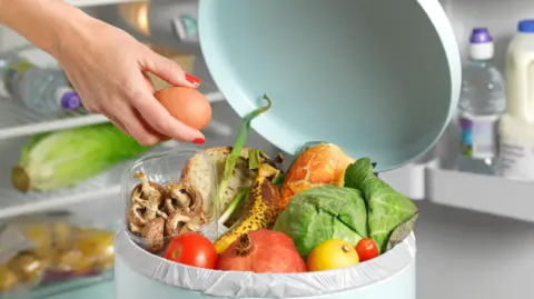 A cylindrical pale blue bin with an assortment of fruit, vegetables and other food inside and hand holding an egg above it