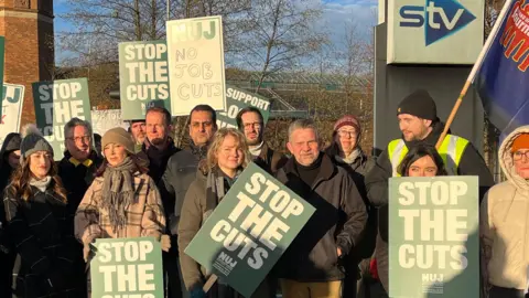 STV staff wearing winter clothing holding placards on a picket line in Glasgow. The signs read "Stop The Cuts" and "No Job Cuts". 