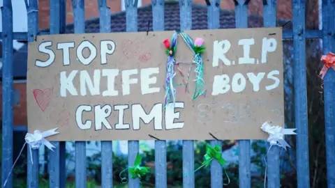 A hand-written cardboard sign hangs on a metal gate. The sign reads Stop Knife Crime. There are flowers and bows hung around the sign.