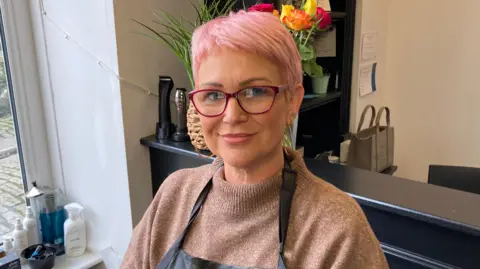 Lisa Steggle, who has short pink hair and is wearing a beige jumper and a black apron and red-rimmed glasses, stands in her hair salon in front of the reception desk, which is black.