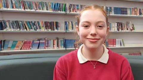 Debbie Tubby/BBC A teenage girl sitting in a library with shelves of books behind her. She has pulled back reddish hair and is starting to smile. She is wearing a white shirt under a red sweatshirt. 