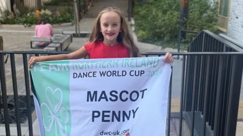 Carrie Maxwell A girl, team mascot Penny pictured with her Team Northern Ireland flag that reads "Mascot Penny" the flag is white with a green stripe down the side and a shamrock. She has blonde hair, is wearing red, along with having red lipstick on.
