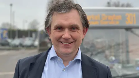 Rob Wilson has long grey hair and is smiling at the camera, standing in front of a bus that has a "market drayton" sign on it. He is wearing a light blue shirt and a dark navy suit jacket.