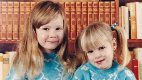 Family of Kirsty Wilkinson Hayley and Kirsty Wilkinson are two girls aged approximately five and three. Hayley, left, has chest-length blonde hair with a long fringe brushed to the side. Kirsty has blonde hair worn in two bunches. They are wearing identical turquoise jumpers with irregular white lines forming patterns on it. They are both smiling at the camera standing in front of bookshelves.