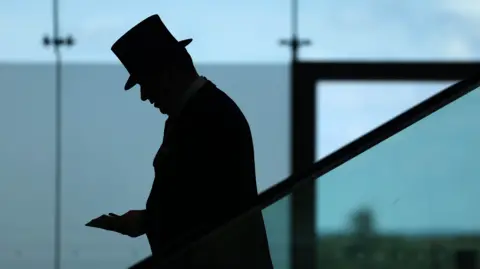 NEIL HALL/EPA-EFE/Shutterstock The silhouette of a man wearing a top hat and suit in front of glass windows at Ascot Racecourse.