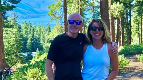 Phil and Anna Brown in a forest with water in the background. They are both smiling at the camera, wearing sunglasses, with each having an arm around the other. They are standing on a pathway through the trees.