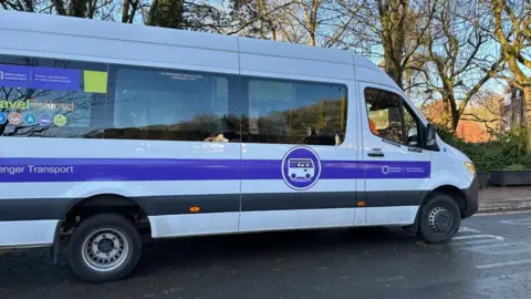 A minibus that has an image of a bus on the side of the vehicle along with Telford & Wrekin council logos. It is on a road and trees are in the background.