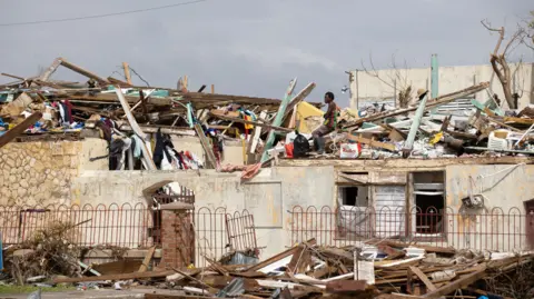 A man searches through the destruction left by Hurricane Melissa in Black River, Jamaica on 11 November. The man is wearing a black, red, green and yellow striped vest and sits on a roof that has been damaged, with piles of planks of wood and other debris sticking out across the rooftops.
