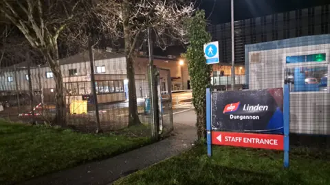 BBC A facility at an industrial park. A sign says Linden Dungannon. Staff entrancement. A fence and trees are in the foreground and a security hut. 