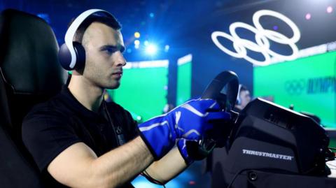  A young man wearing white over-ear headphones sits in front of a videogame controller steering wheel in an arena setting. He wears an expression of concentration as two large screens glow green behind him and a large set of white Olympic rings lit in white hovers over the stage