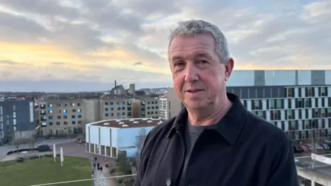 Ben Schofield/BBC Tony Bellot looking directly down the camera, standing outside against a townscape skyline. He has short, greying hair and is wearing a dark jacket over a grey, round-necked top. The sun is setting behind him and on the left of the image is a bright, yellow and orange patch of sky, which is mostly cloudy with the occasional hint of blue. Behind Bellot are modern looking buildings around five or six storeys high, as well as a neat patch of grass in the bottom left of the frame. 