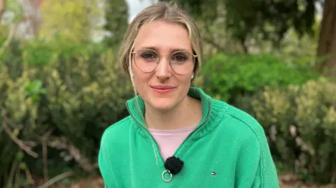 A young white woman with long blonde hair tied back, glasses and a green jumper standing in front of trees and bushes