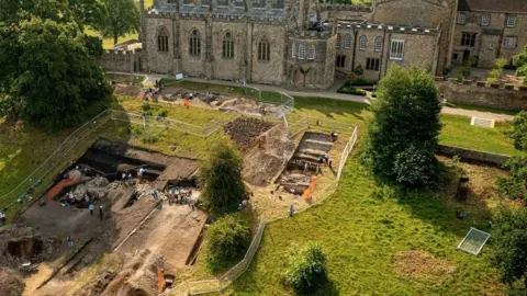 A drone shot of archaeologists digging in fenced off trenches on a grass slope, with Auckland Palace rising behind it. 
