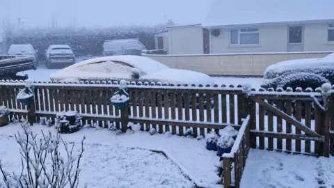 Jon Chilcott Five cars and vans parked on a residential street. Heavy snow has fallen and settled on their roofs and windscreens. There is a front garden fence in the foreground of the picture which is covered in snow. In the distance the sky is grey and foggy with low visibility. 