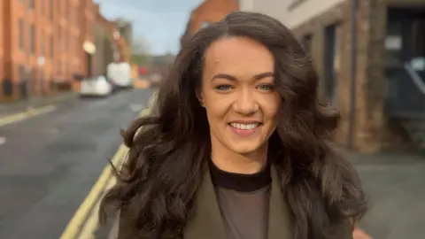 Councillor Charlotte Hill (long dark hair, smiling) in a olive green jacket and dark round neck top