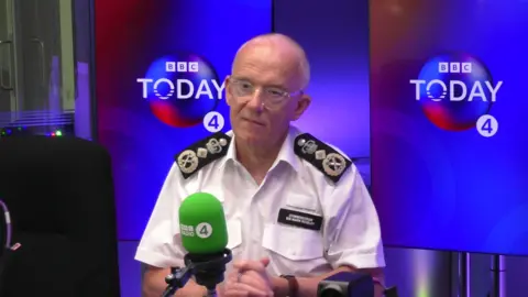 Sir Mark Rowley, wearing a police uniform and glasses, is sitting in the radio studio in front of a green microphone, with the BBC Today programme branding in the background.