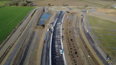 An aerial shot of a road being surfaced. Black asphalt has been laid. There are some works vehicles on the new road, which is in the centre of the shot. On either side of the road, there is a muddy embankment and then green fields. At the top of the picture is a roundabout.