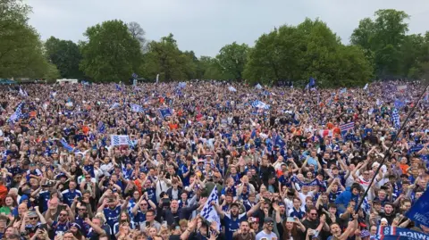 Brenner Woolley/BBC Thousands of Ipswich Town supporters packed together in a park, with banners, scarves, etc. There are mature trees in the background.