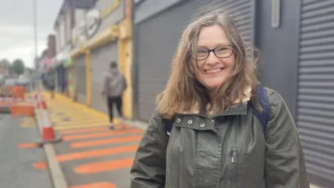 BBC/Claire Hamilton Joy Longshaw, who has shoulder length brown hair and is wearing black rimmed glasses, smiles wearing a green coat while standing on a street. 