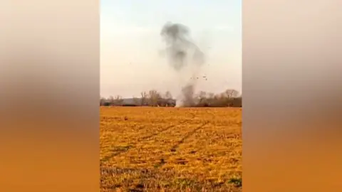 A plume of smoke rising at the other side of a field. Clods of earth are shooting into the sky.