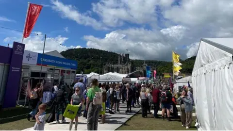 Crowds of people at this year's Eisteddfod. There are stalls on either side of the path in white tents, with Margam Castle behind.