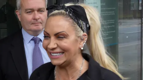 Laura Foster/BBC Jodie Marsh squinting while giving a big smile. She has long blonde hair in a ponytail, held in place by a black hair tie. She is wearing black in front of her lawyer, who is wearing a suit.
