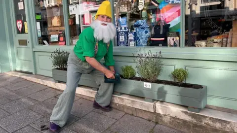 Ellen Knight/BBC Councillor Sue Sealy, wearing a yellow pointed hat, false white beard, a green top, grey braces and green trousers, is leaning over a light green planter. The planter has rosemary and lavender in it, and is outside a shop front painted in the same colour. 