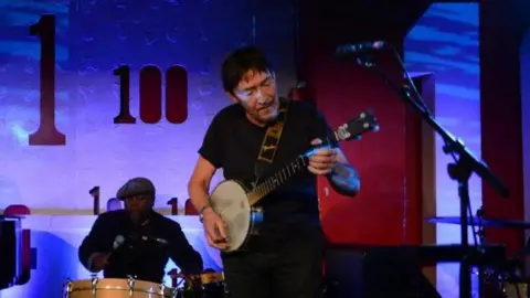 Mark Allen/BBC Chris Rea plays a banjo-type instrument. He is dressed all in black, and behind him a man hits a large drum.
