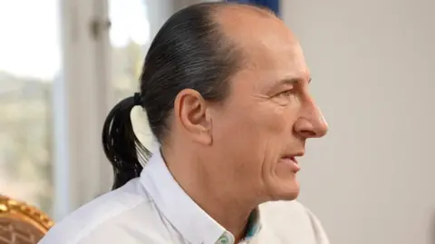 A man in a white shirt with a black pony tail is pictured side-on