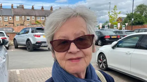 A woman with short grey hair and large sunglasses and a blue scarf looks towards the camera.