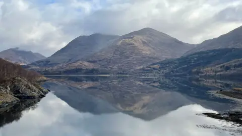 Mountains, their peaks just covered in low cloud, reflected in a loch. The sky is both grey and blue.
