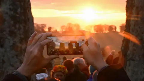 Reuters A person taking a photo of the sunrise during the Spring Equinox at Stonehenge. The image shows a person holding their phone horizontally while they take a picture. The stone circle can be seen on the screen of their phone. 