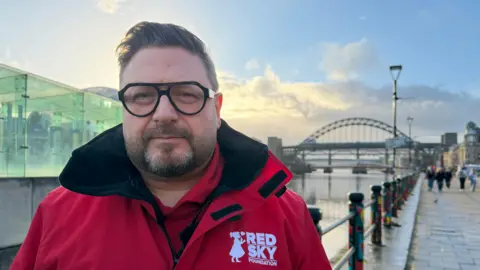 Sergio Petrucci is standing on the Newcastle Quayside with the Tyne Bridge behind him. He has short brown hair, glasses and is wearing a red 'Red Sky Foundation' branded jacket.
