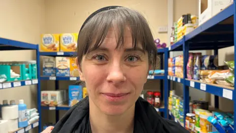 A brown haired woman, with a fringe and blue eyes looking into the camera.
She is wearing a black scarf and is stood in front of shelves of tinned food.