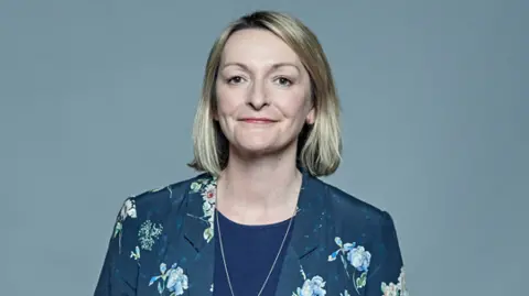 PA Media Jess Morden head shot. She has a blonde bob and smiles at the camera. She has a blue top with a silver chain and a blue and flowery blazer on.