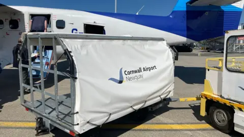 A baggage trolley with Cornwall Airport Newquay on the side of it, in front of a plane