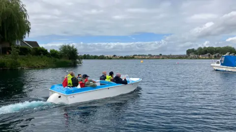 A blue and white speedboat is being driven across a large lake. There are five people in the boat, three in the front and two in the back. They all have life jackets on. The boat has a red flag on the back of it. 