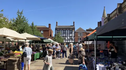 A pedestrianised street with market stalls and a large number of people walking around. There are buildings in the background, with a black and white building being the most prominent. The scene is set against a blue sky.