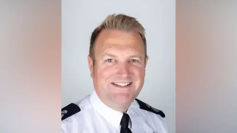 Cambridgeshire Fire and Rescue Service A headshot of Stuart Smith who is smiling straight at the camera while wearing a smart uniform - a white shirt with a black tie.