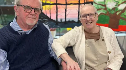 Sally and Kevin are looking at the camera, holding hands and smiling. Kevin is wearing a blue and white stripped shirt and a navy knitted tank top. Sally is wearing a cream cardigan, brown t-shirt and light brown dungarees. Both have short white hair and glasses.