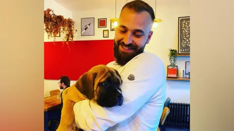 A man with short brown hair is hugging a puppy dog he is holding in his hands. He has a thick brown beard and is smiling. He is wearing a white sweater and he is in a cafe.