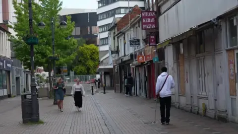 一对妇女走在斯温顿的一条街道上，前面一家用木板封住的商店前面，右侧有一名身穿白色连帽衫、拄着拐杖的男子。