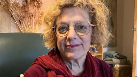 A woman wearing round glasses and red lipstick smiles at the camera. She's also wearing dangly earrings, a velvet red top and scarf. Behind her is an example of her husband's art work on the wall. 