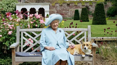 Historic England Queen Elizabeth the Queen Mother sits alongside a Corgi dog on a bench in the Walmer Castle gardens