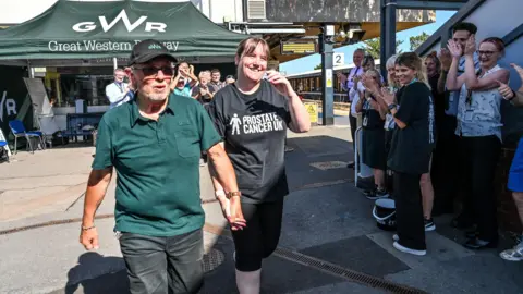 An older man wearing a dark green polo shirt and black Great Western Railway cap holds the hand of a woman with brown hair wearing a black Prostate Cancer UK T-shirt as they walk down a railway platform. Other members of railway staff can be seen smiling and clapping. There is a dark green Great Western Railway marquee behind them.