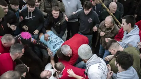 Several men watching and reaching down to get a leather tube from a man who is on the floor. They are dressed in hoodies, black tops.