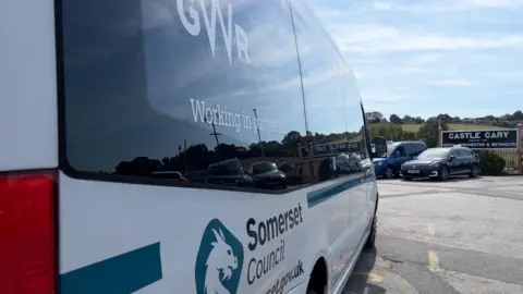 A minibus stretches into the centre of the frame from the left hand side of the photo. It says 'Somerset Council' on it, with 'working in partnership with GWR' above. A black railway sign for Castle Cary is in the distance.