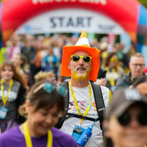 Elaine Livingstone A man wearing an orange and white traffic cone on his head at the start of the Kiltwalk. He is also wearing a yellow lanyard and has dyed his beard yellow