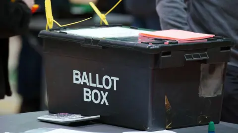 The image shows a sealed black ballot box on a table. A grey calculator is on the table next to it along with a green rubber thimble. A pad of orange paper is on top of the box.